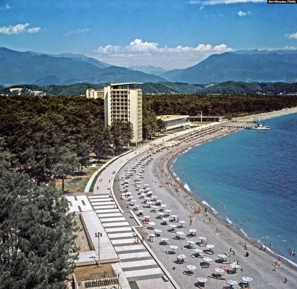 A colour photograph of the beach at Pitsunda, with a tall hotel among pines and the mountains beyond